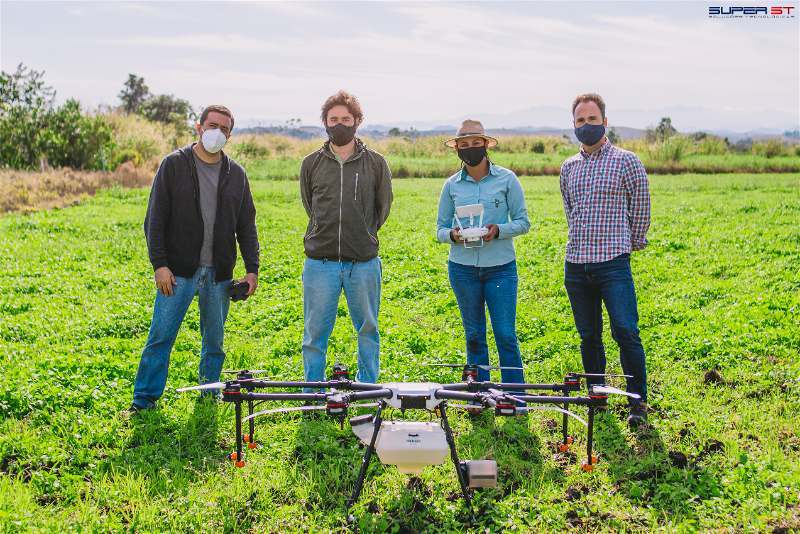 Imagem de quatro profissionais da agricultura em um campo aberto, observando um drone em funcionamento